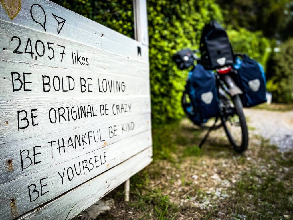 Schild mit Text:
Be Bold Be Lovin
Be Original Be Crazy
Be Thankful Be Kind
Be Yourself
Im Hintergrund Oliver Baiers bepacktes Reiserad